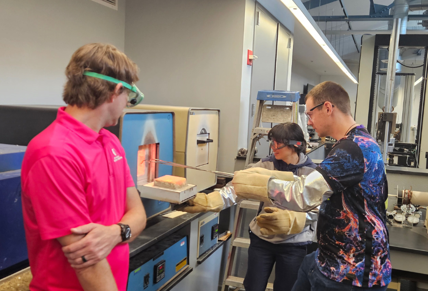 Three people in a laboratory observe a sample being pulled from a high-temperature furnace in use